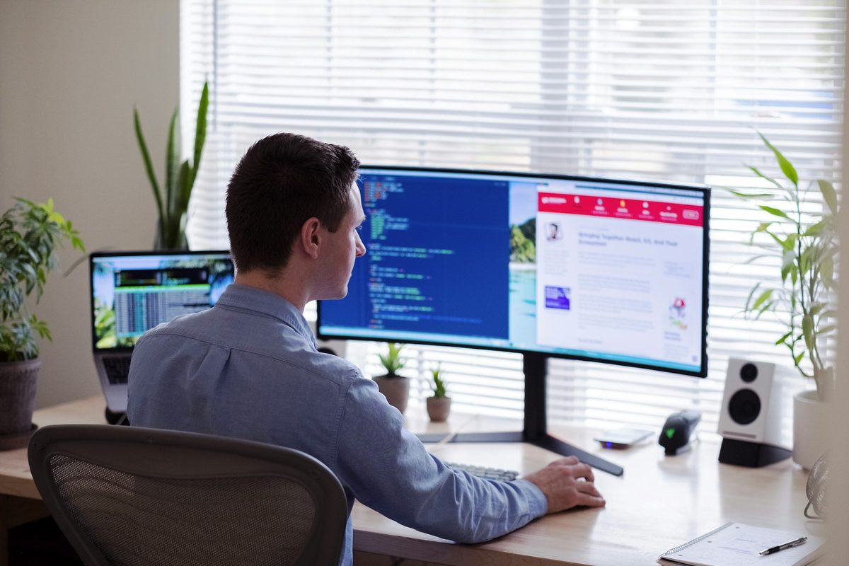 Someone working at a bright, plant-filled desk with a wide monitor, headphones on, focused on their screen.